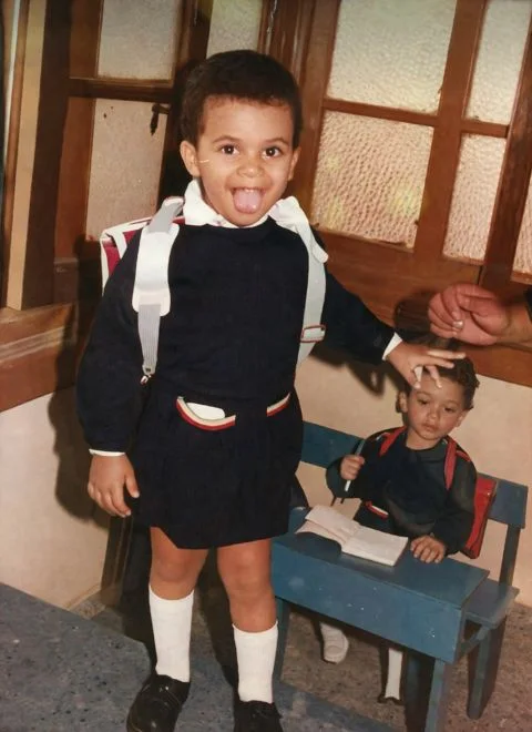 Peter GORGY as a young child on his first day of pre-school, grinning with his tongue out in a navy school uniform and backpack, with a classmate seated at a desk behind him